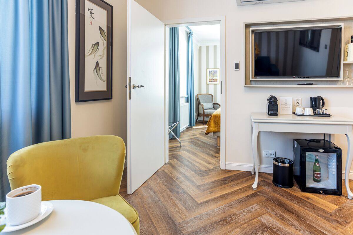 A hotel room with a yellow armchair, a white table with a cup, a TV, a minibar, and a door leading to another room.