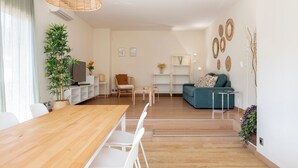 Un salon moderne avec une table à manger en bois, des chaises blanches, un canapé bleu et une petite table basse. On y trouve une plante en pot et un téléviseur posé sur un meuble.