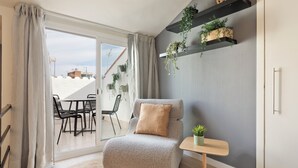 Un salon moderne avec un fauteuil gris, une petite table d’appoint en bois ornée d’une plante, et une vue sur un balcon aménagé avec du mobilier d’extérieur.