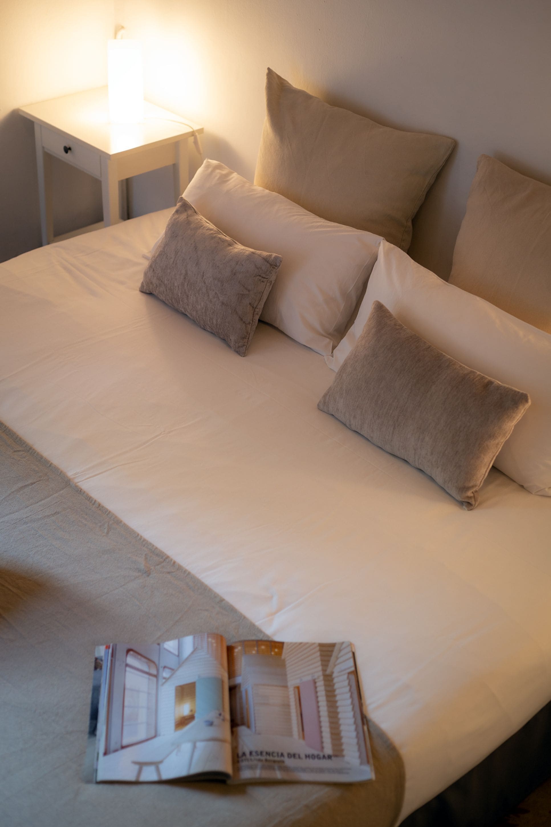 A neatly made bed with a magazine on it, a bedside lamp, and a nightstand.