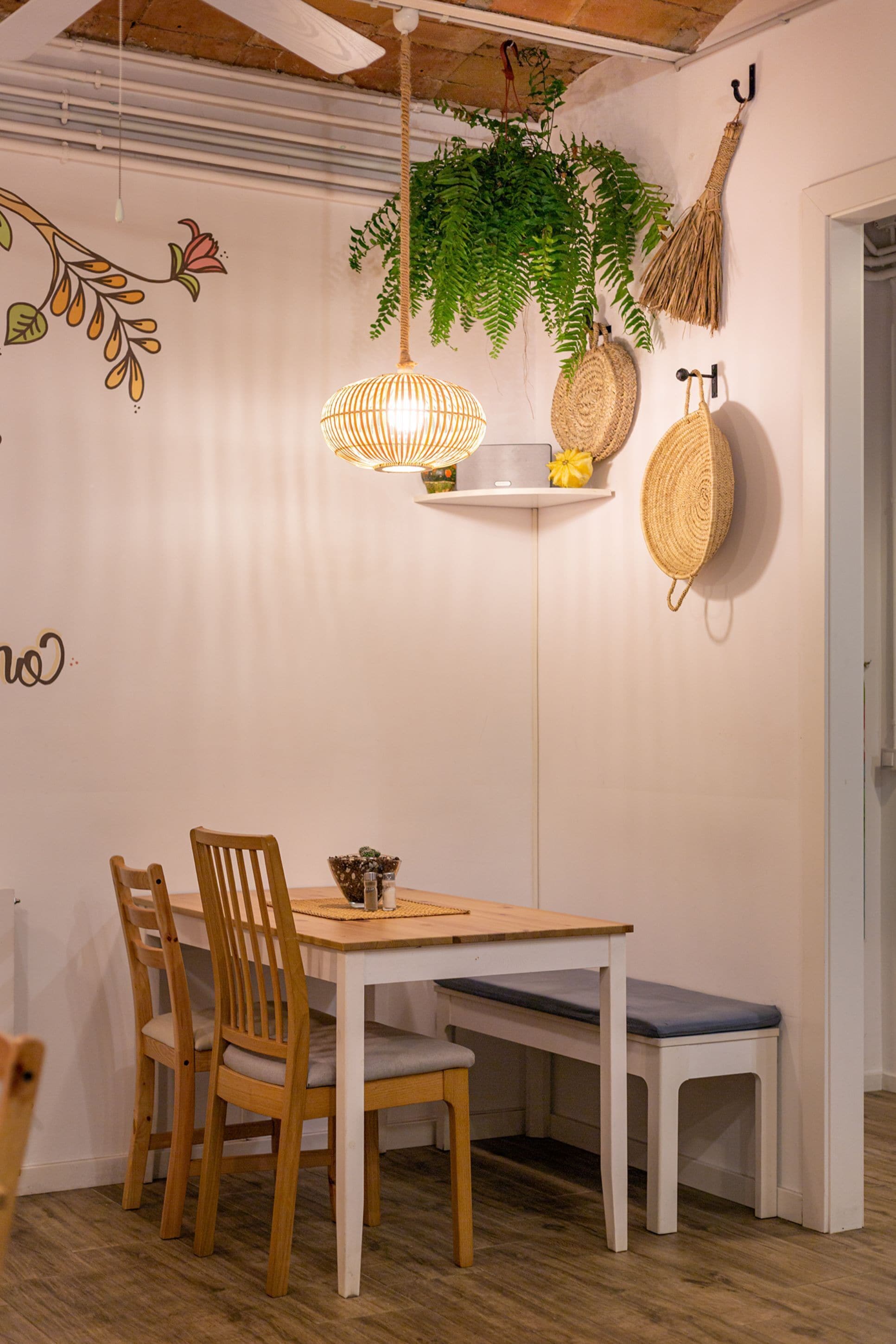 Un comedor con una mesa de madera, cuatro sillas, un banco, una planta colgante, una cesta tejida y una lámpara de pared.