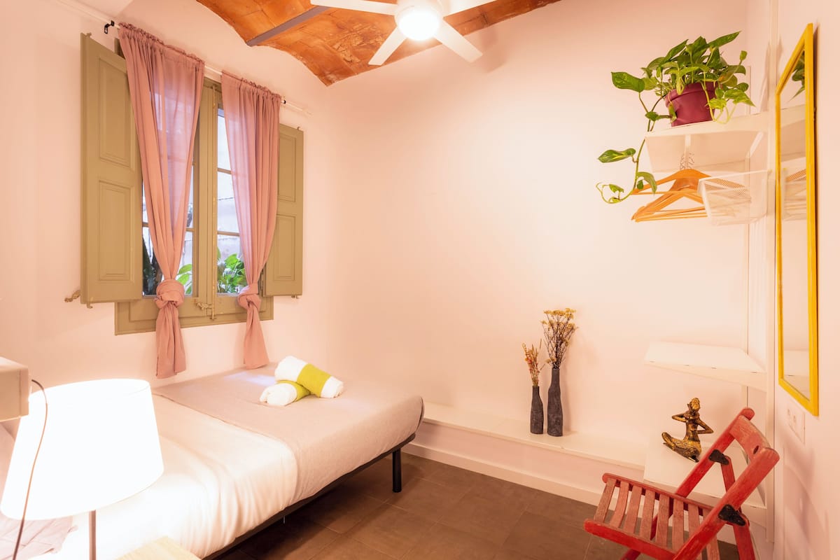 Ein Schlafzimmer mit Bett, Sessel, Schreibtisch, einem Fenster mit Vorhängen, einem Deckenventilator, einem Regal mit Pflanze und einem Spiegel.