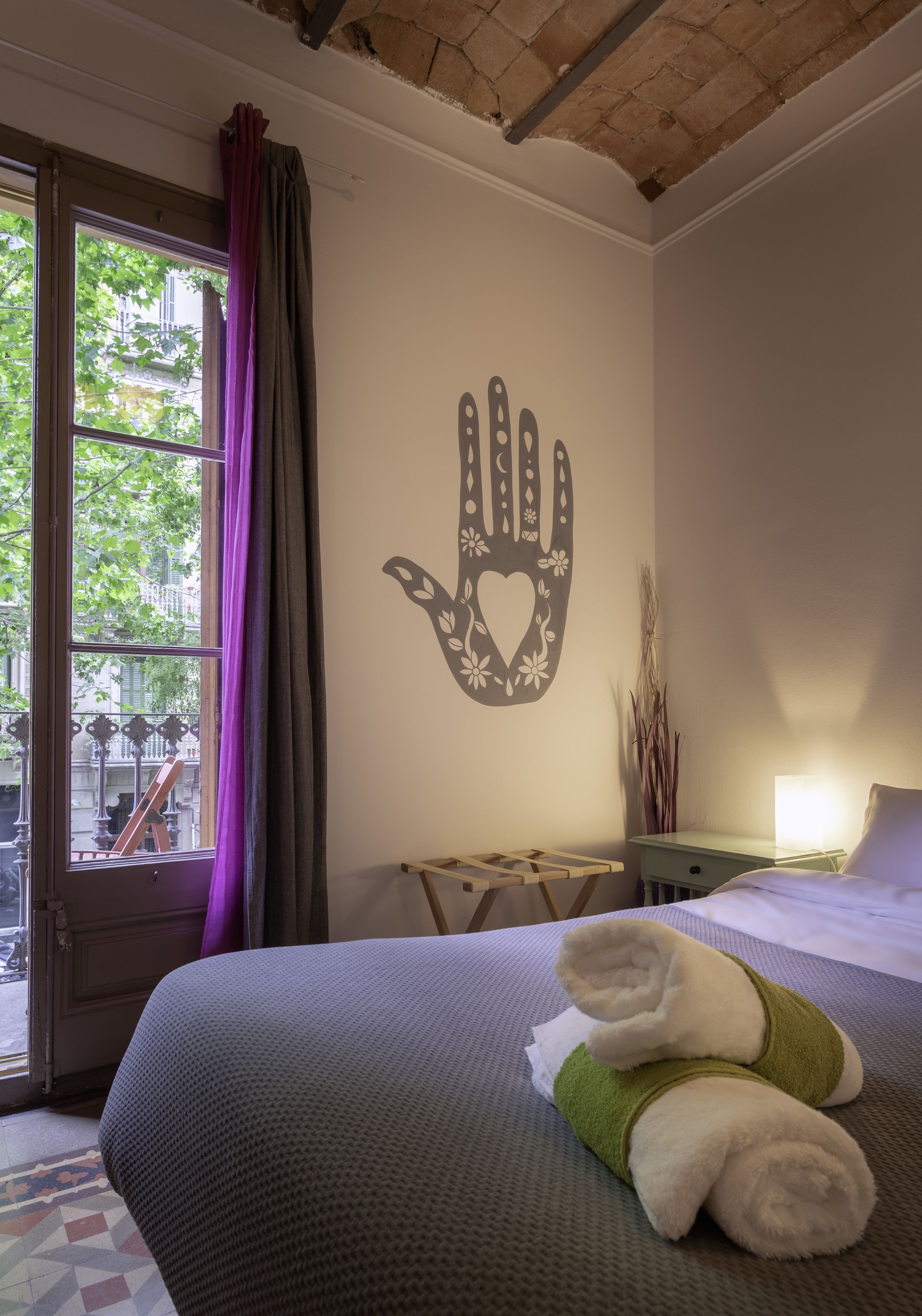 Un dormitorio con una cama, un adorno en la pared con forma de mano, una ventana con cortinas y una mesita de noche con lámpara.