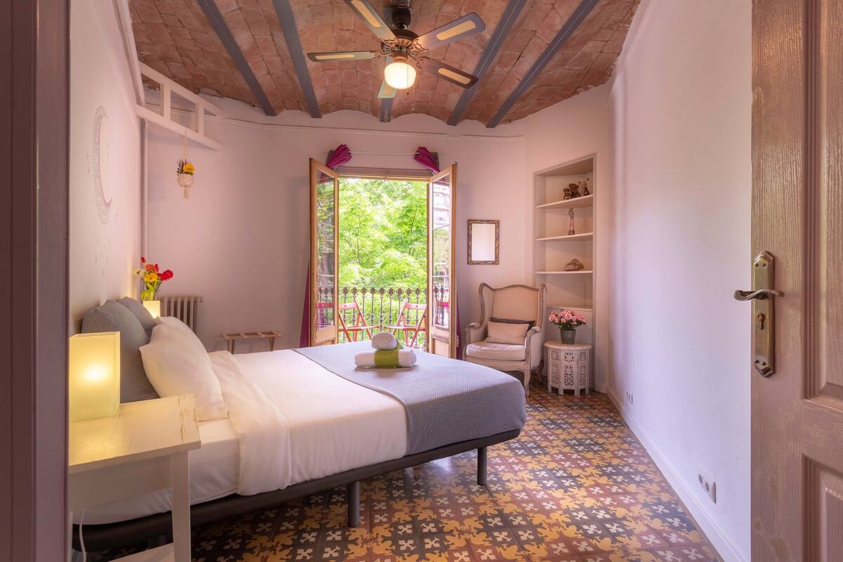 Ein Schlafzimmer mit Bett, Schreibtisch, Stuhl, Heizkörper, Fenster mit Blick auf Grünflächen und Deckenventilator.