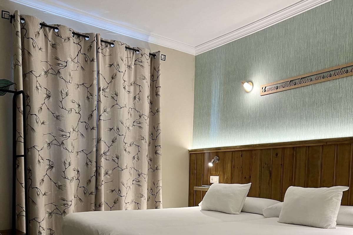 Habitació d'hotel amb un llit gran, un capçal de fusta, una taula de nit i una cortina amb motius de fulles.