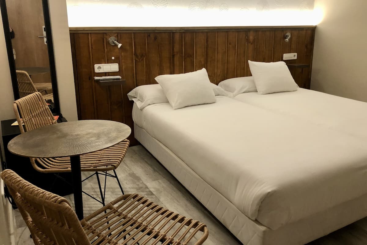 Una habitació d'hotel amb un llit gran, dos coixins blancs, una taula rodona petita i cadires de vimet.