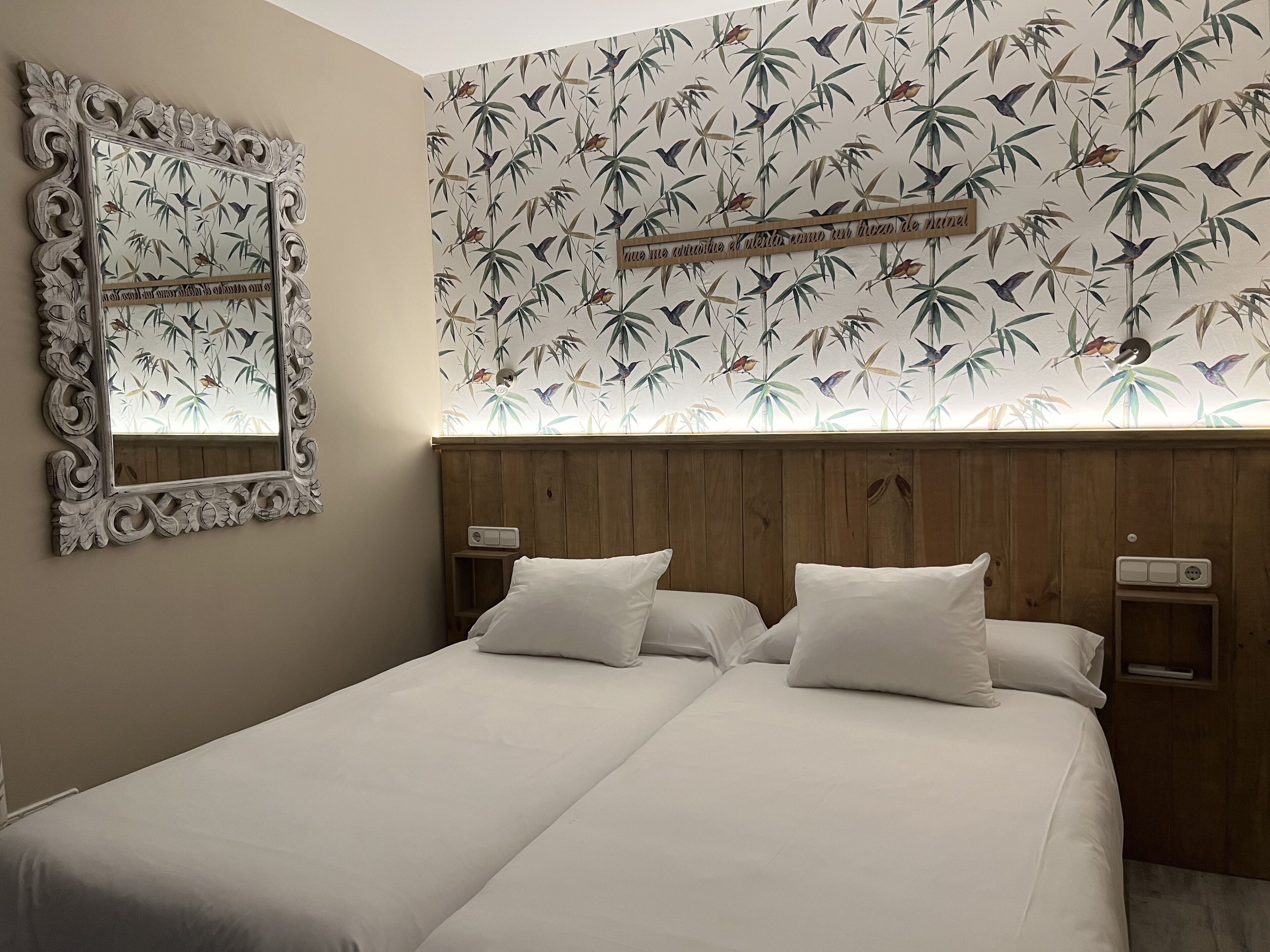 Un lit double avec du linge de lit blanc, dans une pièce aux murs lambrissés et recouverte d’un papier peint à motifs de feuilles de bambou.