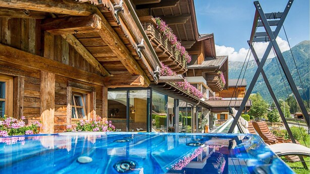 Una cabaña de madera con piscina, balcones adornados con flores y un paisaje de montaña al fondo.