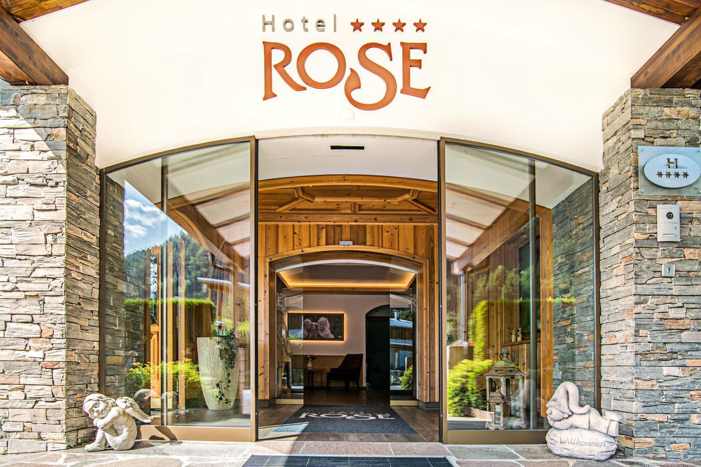 A entrada do Hotel Rose, com fachada de pedra, portas de vidro e arco de madeira.