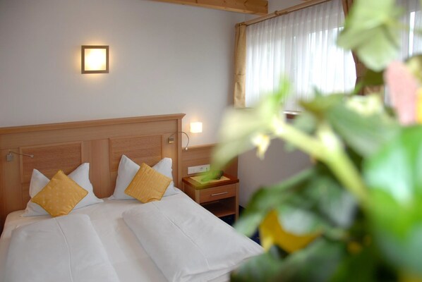 A hotel room with a wooden headboard, two beds with white linens and yellow pillows, a bedside table with a green object, and a wall-mounted light.