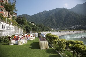 Montaje de boda con una mesa y sillas en un área cubierta de césped, un letrero que dice 'Divine Resort', y un río con montañas al fondo.