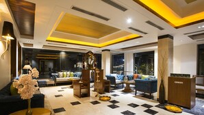 Eine moderne Lobby mit Schachbrettmuster-Boden, einer zentralen Sitzecke mit Sofas und Sesseln, einer dekorativen Säule und einem Empfangsbereich.