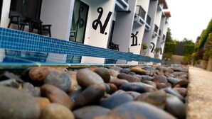 A hotel entrance with a tiled pathway leading to the entrance, a series of columns with signage, and a seating area visible in the background. - Alphabeto Resort (Rawai)