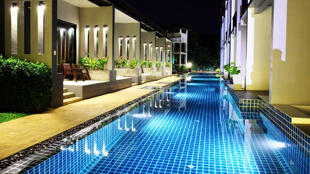 A swimming pool with a tiled edge, adjacent to a building with a covered seating area and potted plants. - Alphabeto Resort (Rawai)