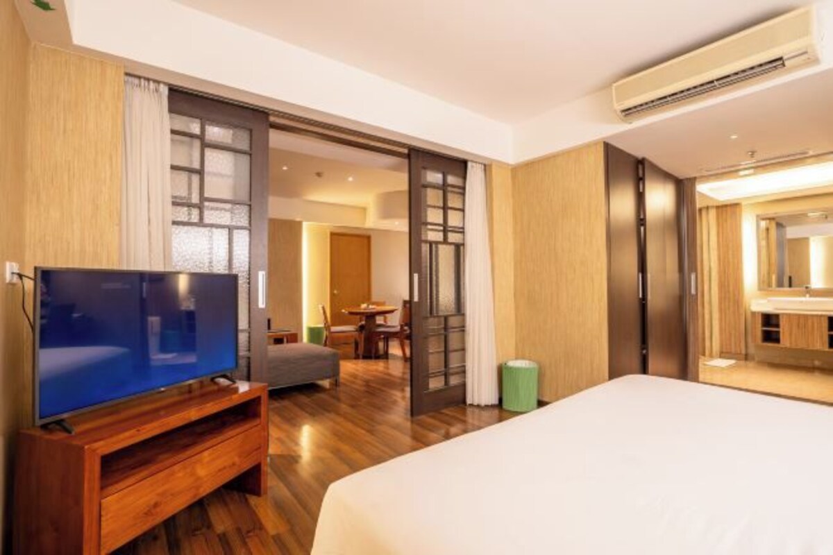 Quarto de hotel com cama, televisão sobre suporte de madeira, área de refeições e banheiro com espelho.