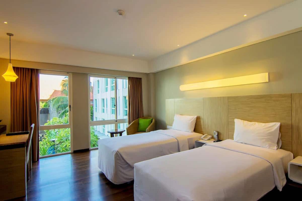 A hotel room with two beds, a desk, a chair, a phone, and a window with a view of buildings and greenery.