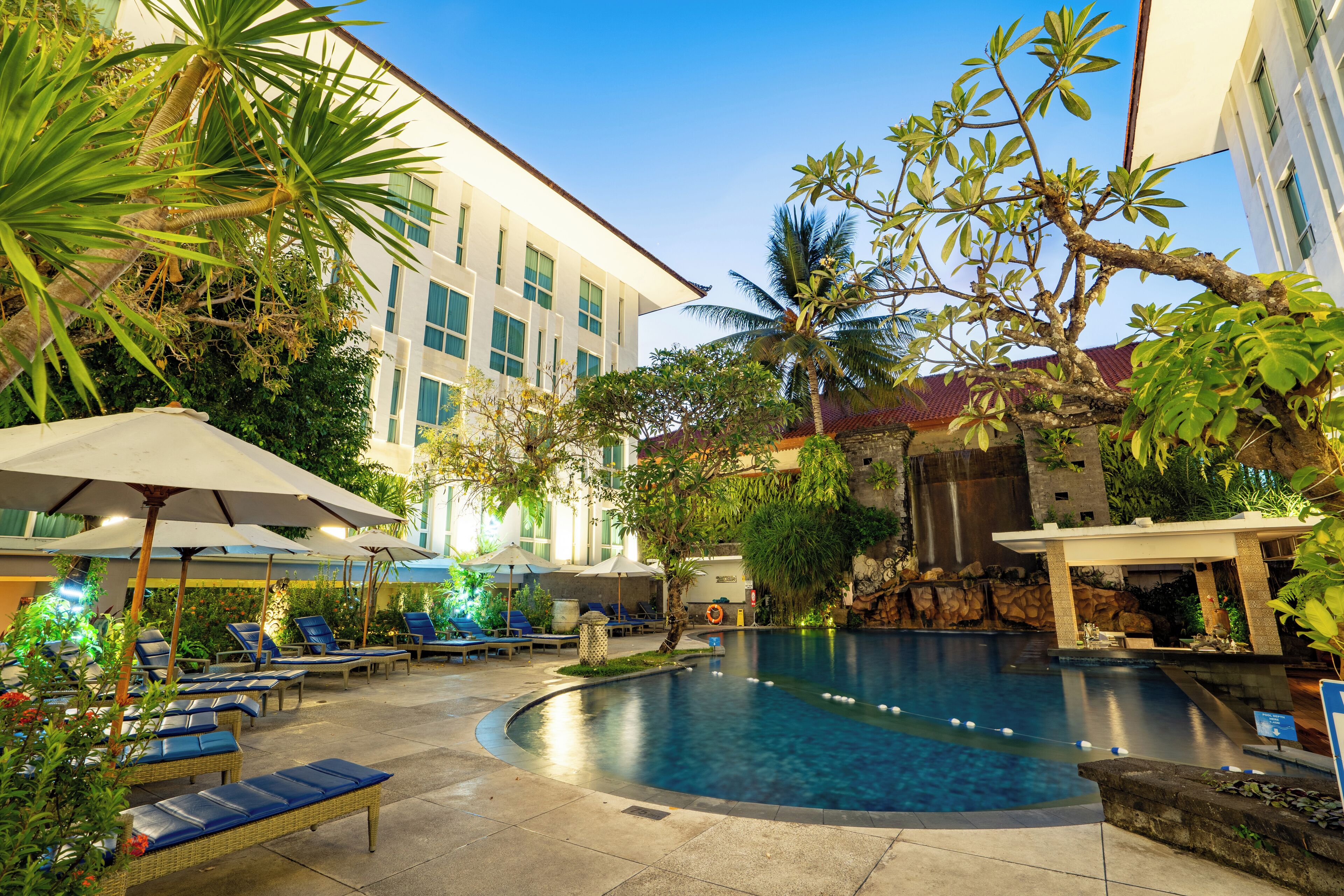 A pool area with lounge chairs and umbrellas, surrounded by greenery and a building with a waterfall feature.
