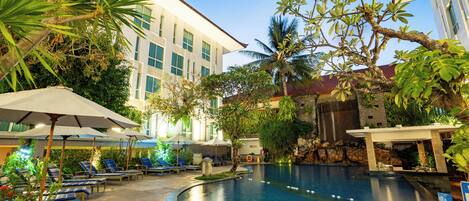 A pool area with lounge chairs and umbrellas, surrounded by greenery and a building with a waterfall feature.