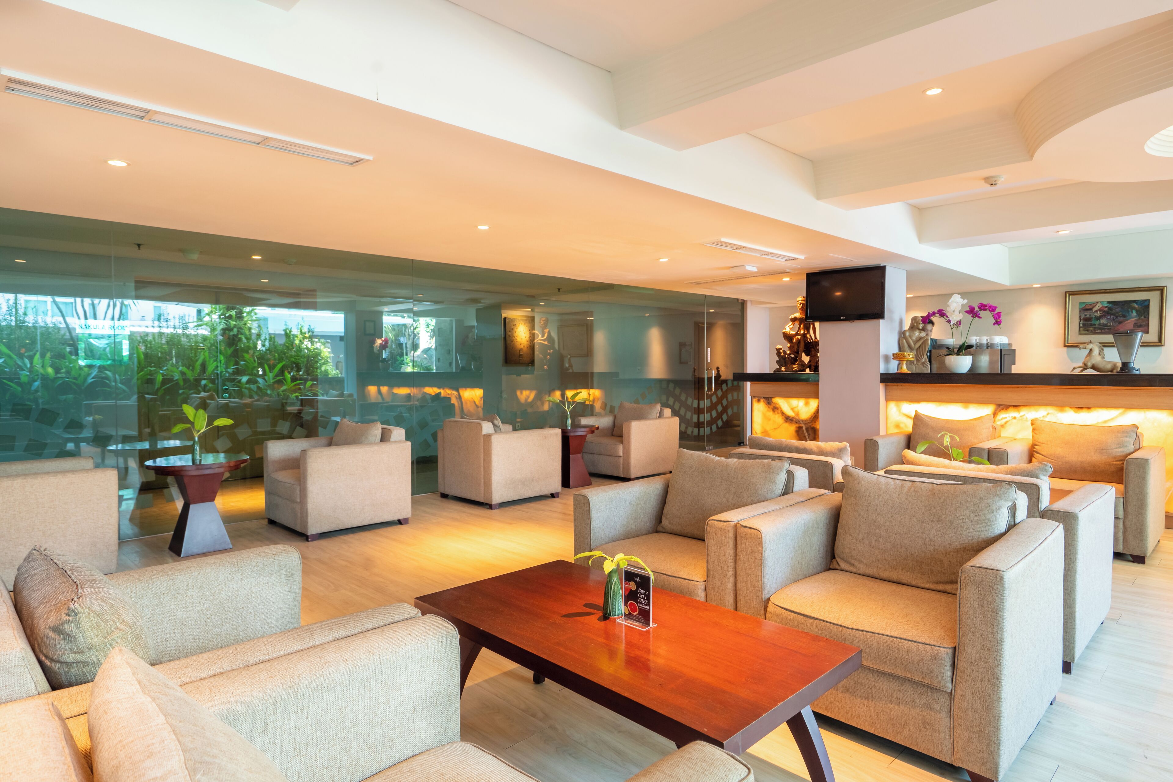 A spacious living area with multiple sofas and a coffee table, a television mounted on the wall, and a large window offering a view of greenery outside.
