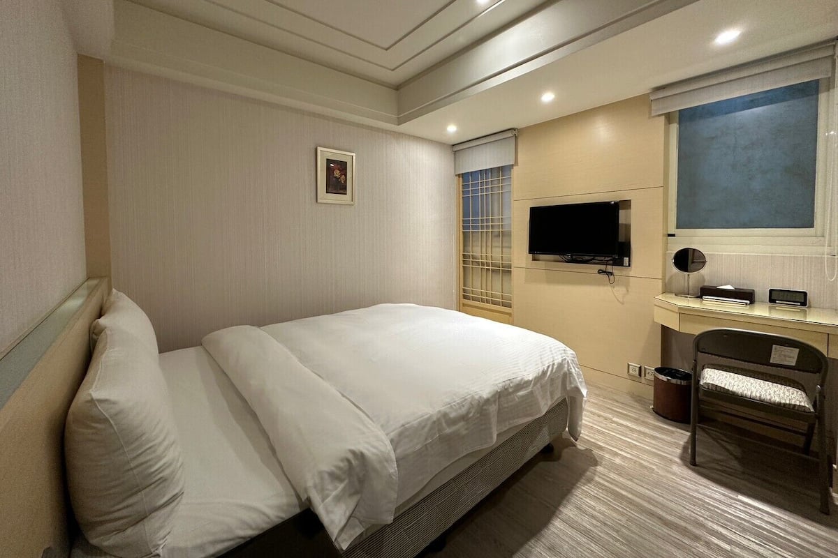 A hotel room with a large bed, a desk with a TV, and a window with blinds.
