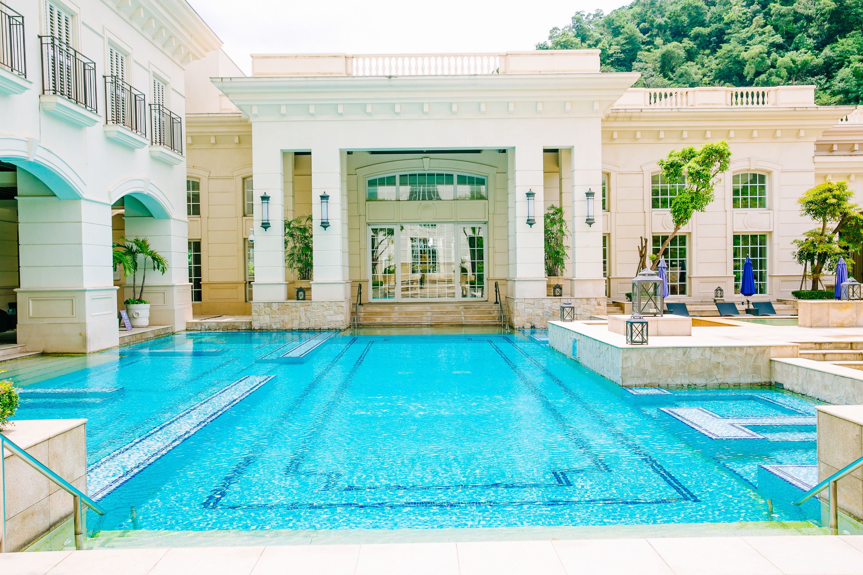 A large indoor swimming pool with clear blue water, surrounded by a building with classical architecture, including columns and a grand entrance.