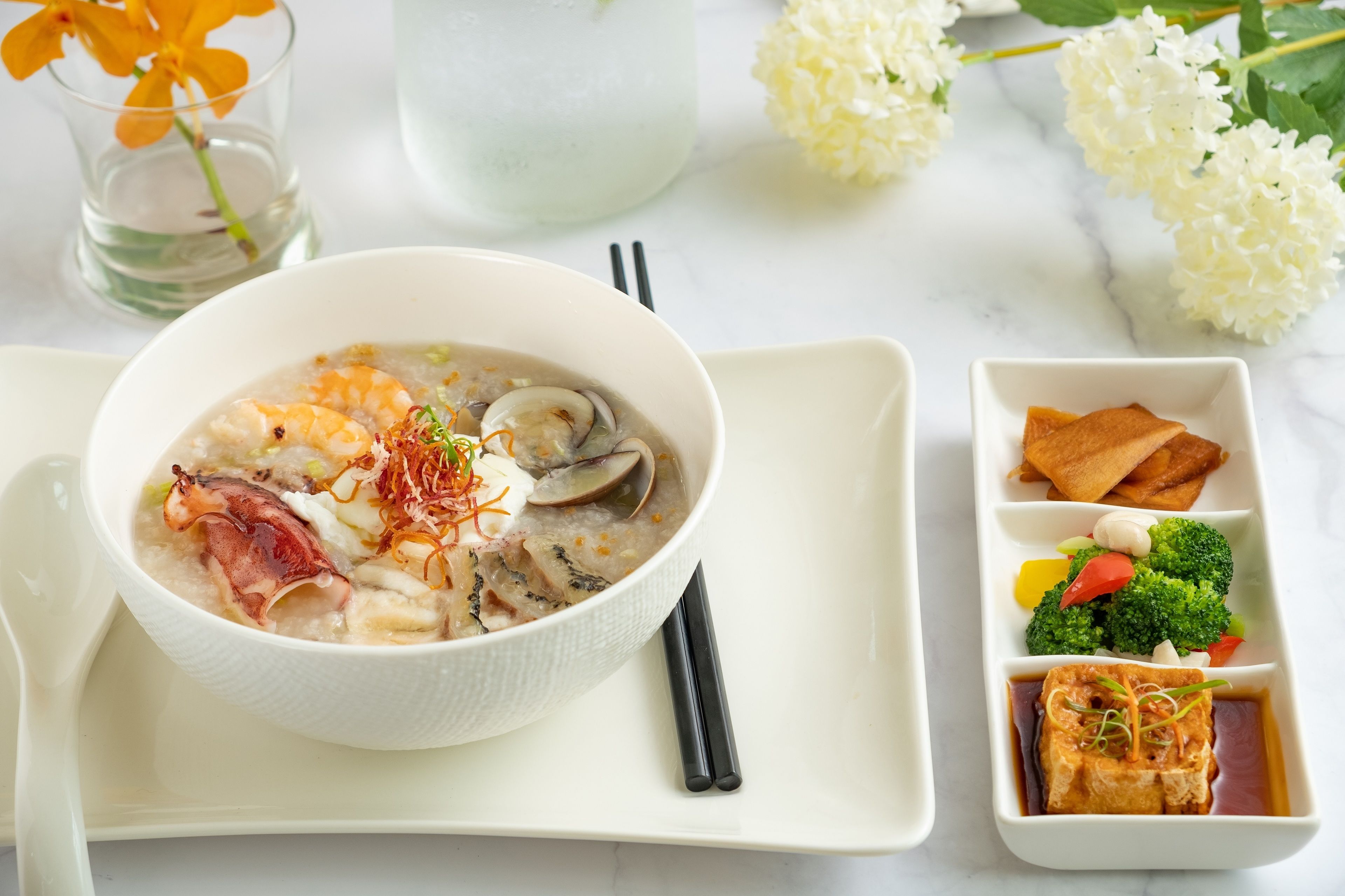 A bowl of soup with various ingredients, a plate with tofu and vegetables, and a glass of water with flowers.