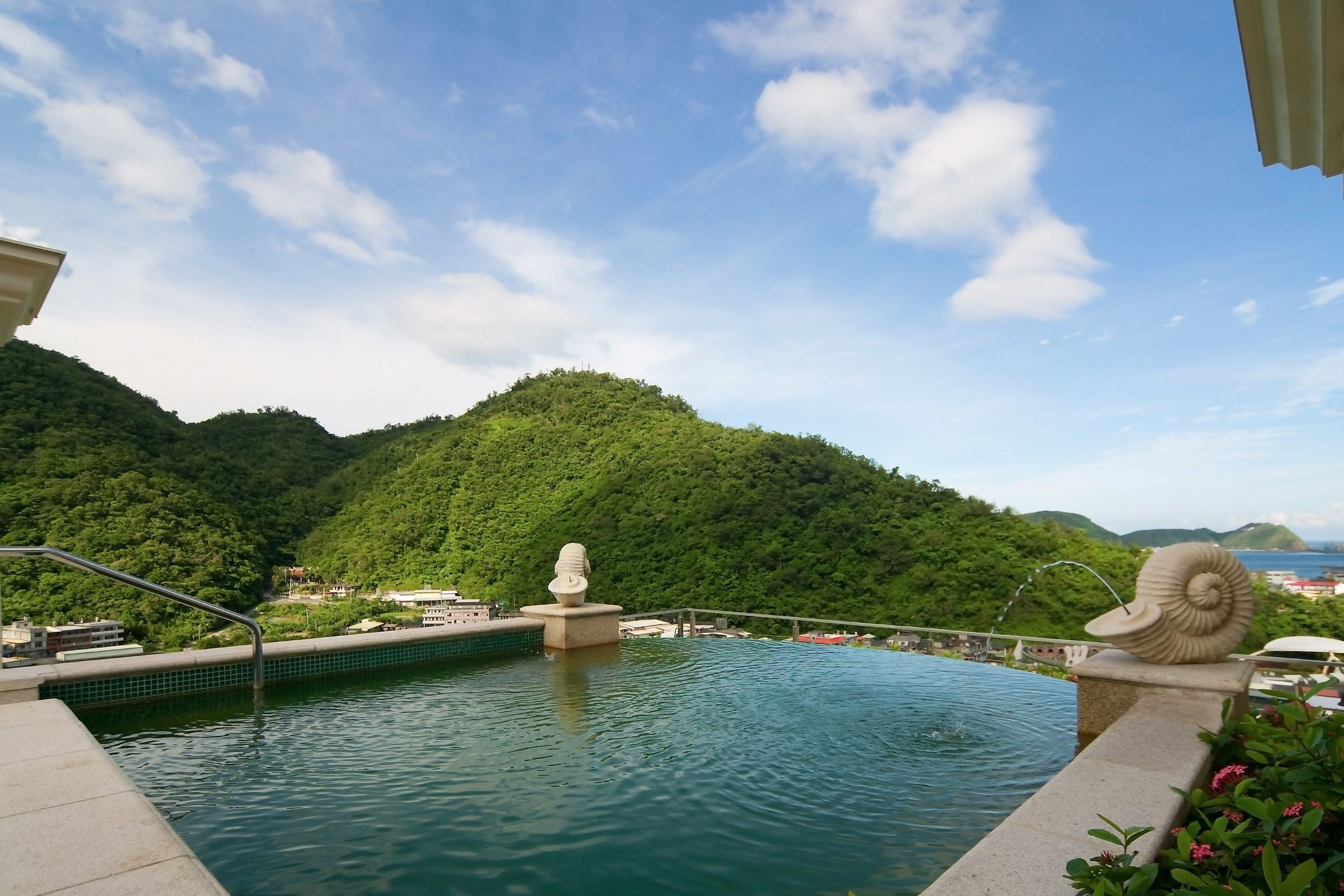 A pool with a statue and a shell decoration, surrounded by greenery and hills.