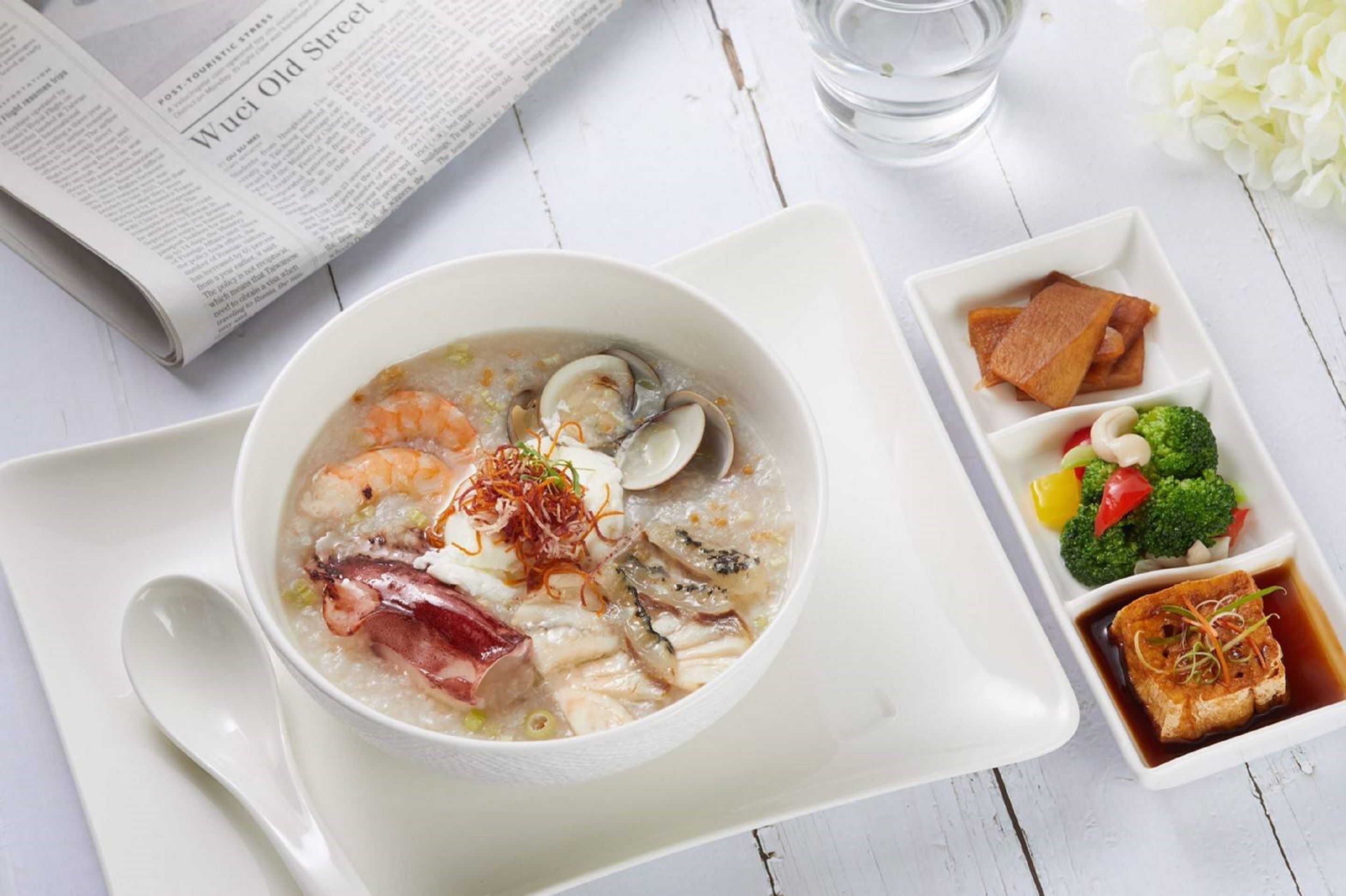A bowl of soup with various ingredients, a newspaper, a glass of water, and a plate with vegetables and tofu.