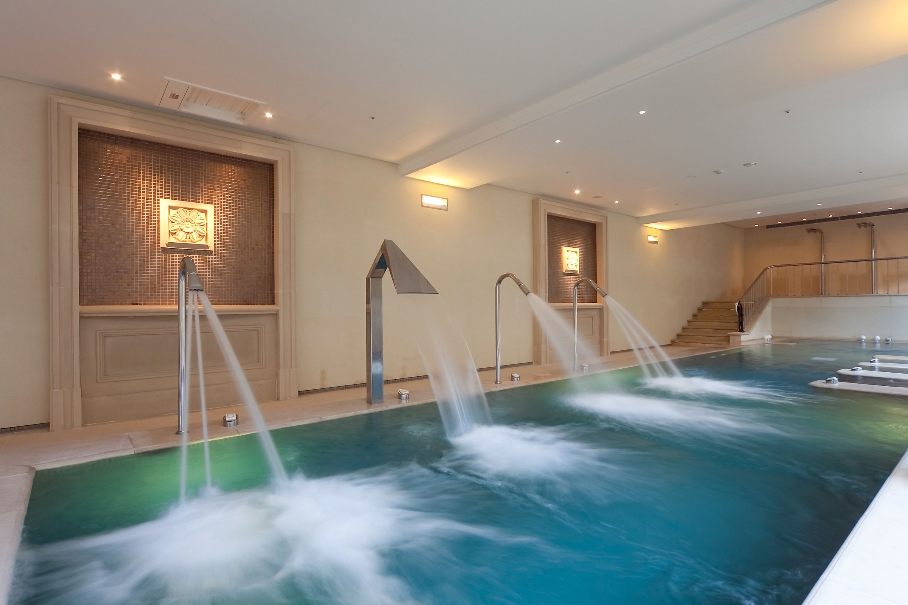 A large indoor swimming pool with multiple water jets, a tiled wall with a decorative panel, and a staircase leading to an upper level.