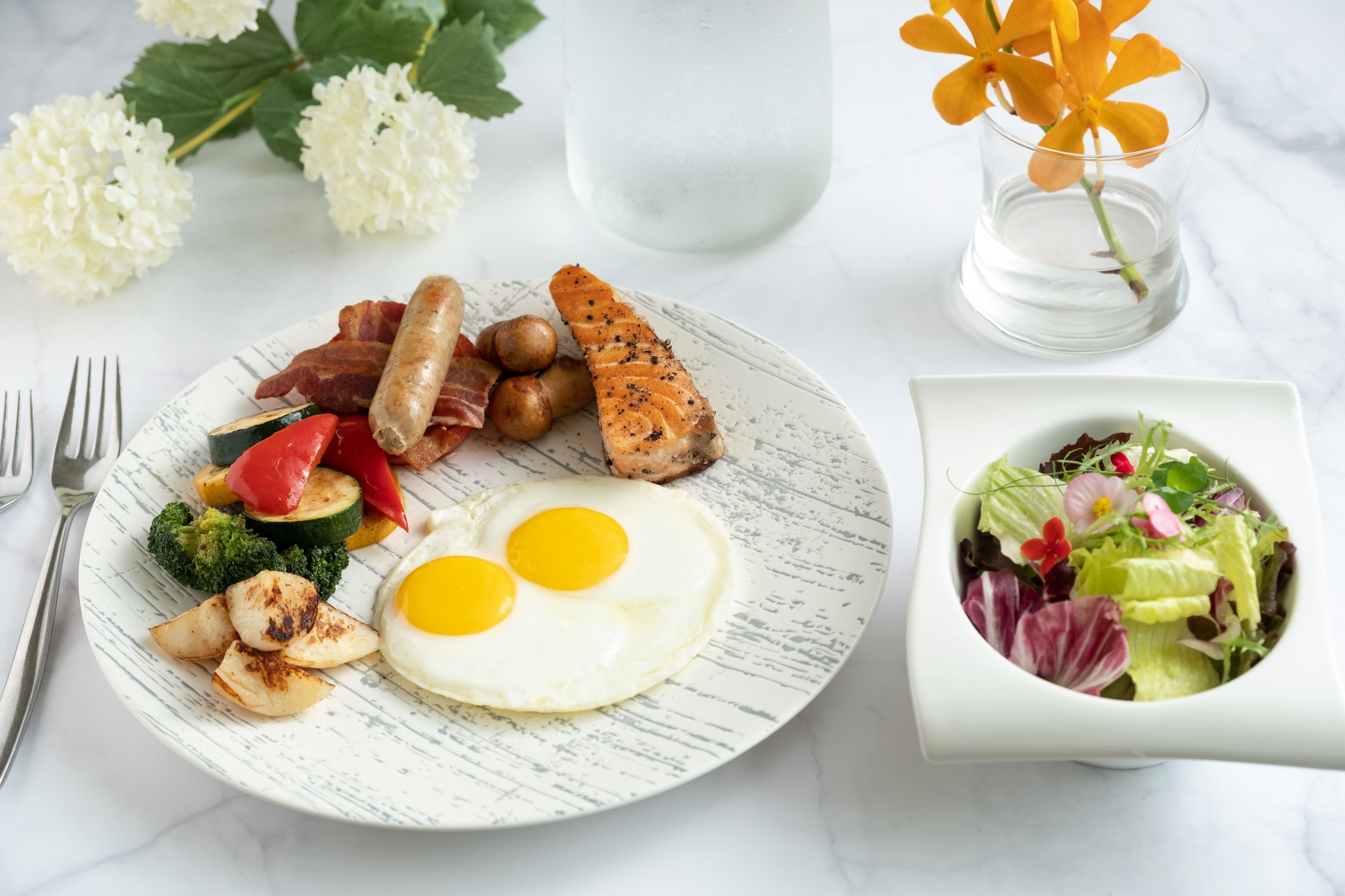 A breakfast plate with eggs, bacon, sausage, and vegetables, a glass of water, and a bowl of salad.