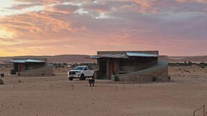 A desert landscape with two modern buildings, one with a sloped roof and another with a flat roof, both featuring brick construction and a small attached structure. A white pickup truck is parked in front of the buildings. - Sossus Oasis Camp Site (Sesriem)