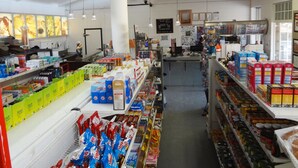 A grocery store interior with shelves stocked with various products, a checkout counter, and a cashier. - Sossus Oasis Camp Site (Sesriem)