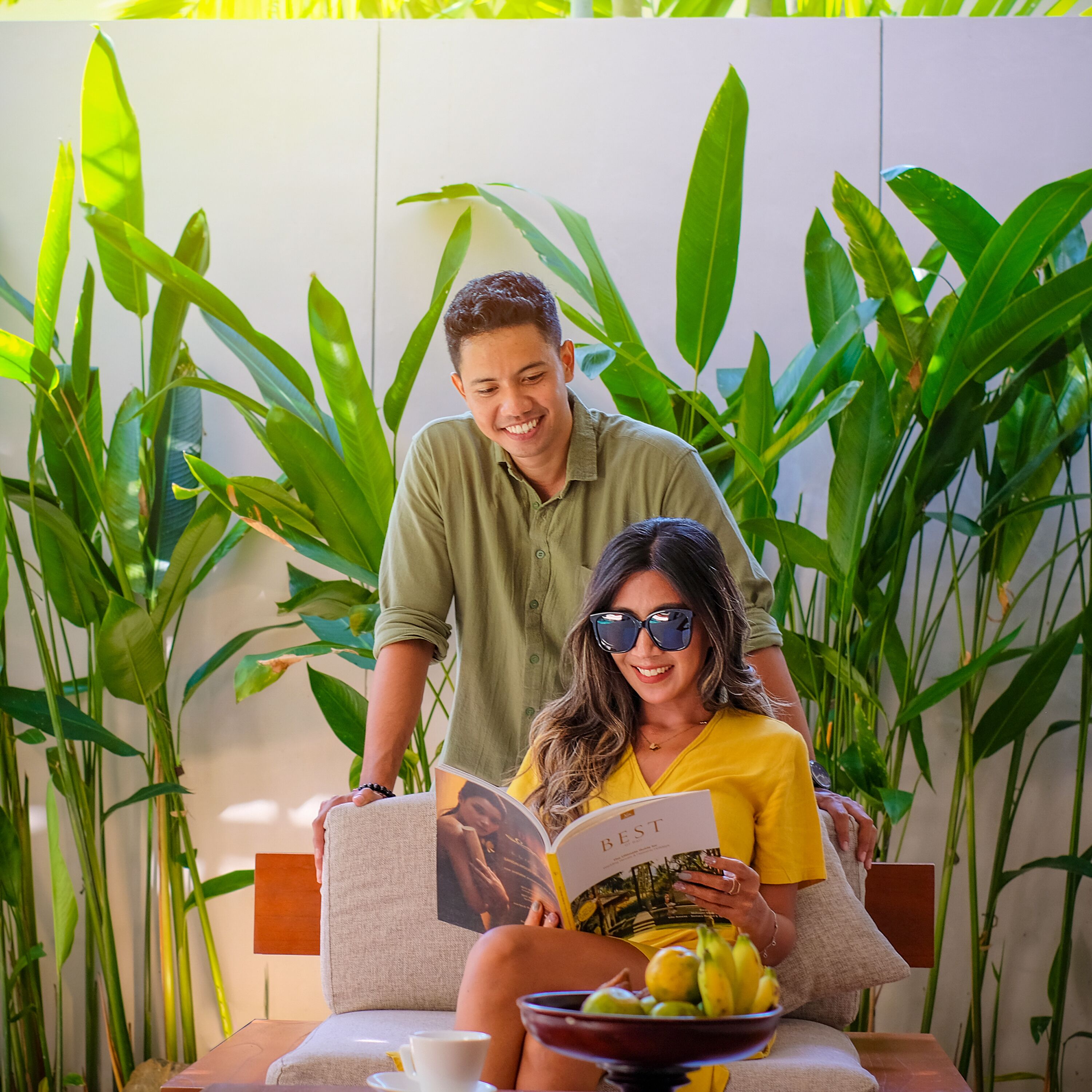 A couple seated on a sofa with a bowl of fruit and a book, surrounded by green plants.