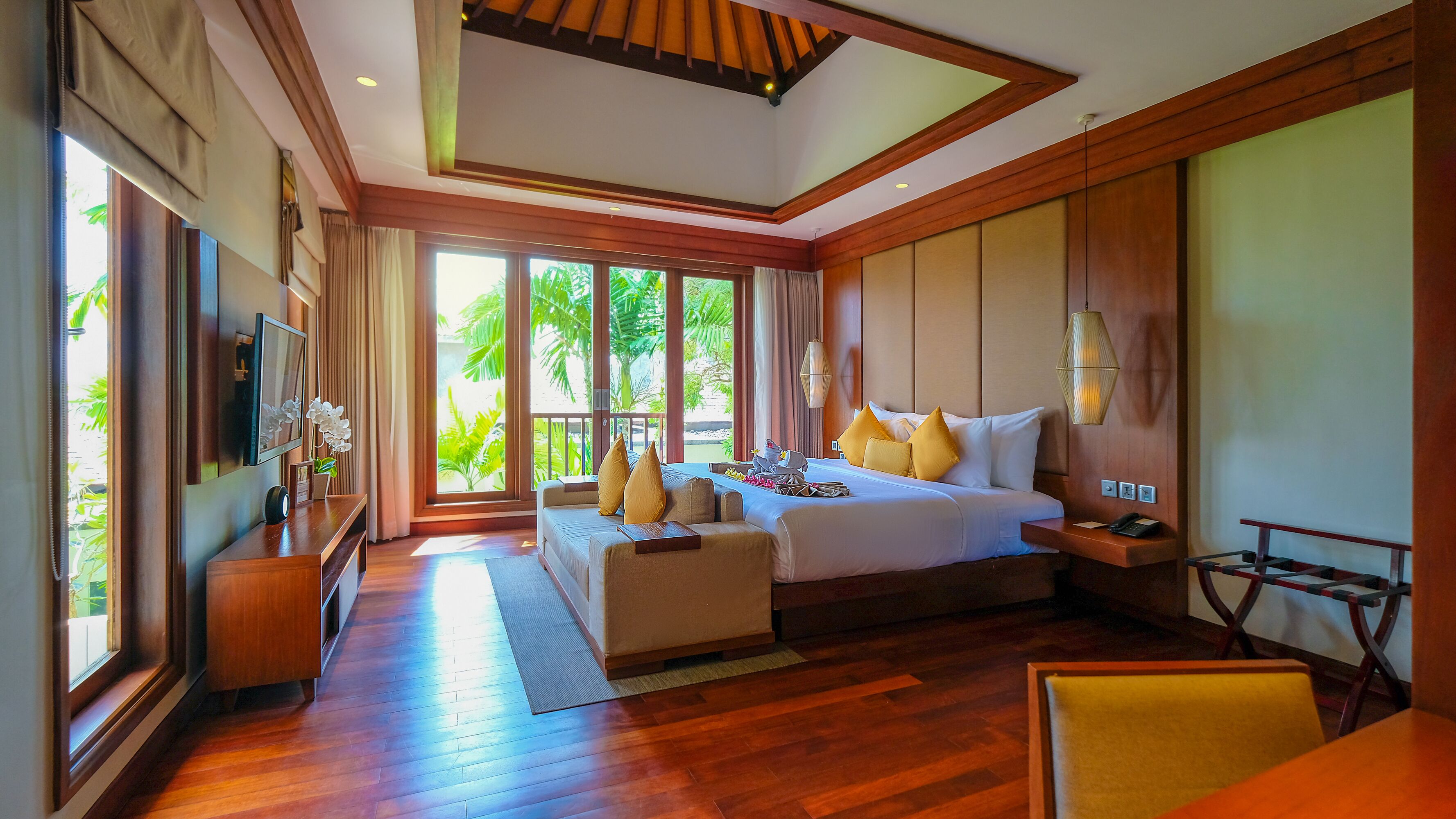A spacious bedroom with a large bed, a sofa, and a desk. There is a window with a view of greenery outside.