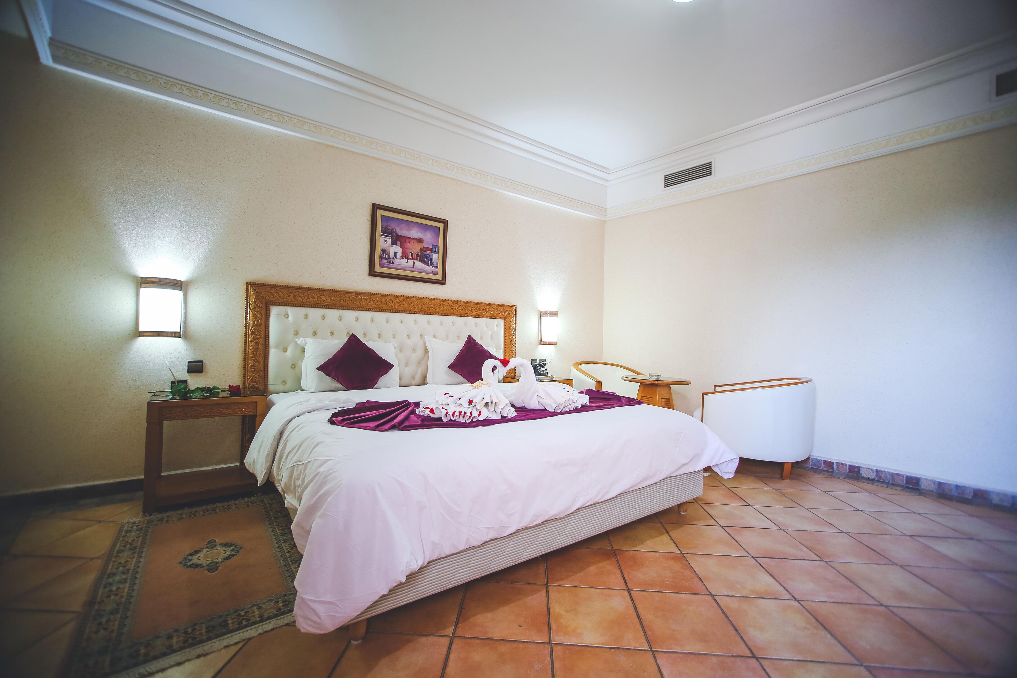 A hotel room with a large bed, a nightstand, a lamp, a wall-mounted picture, and a tiled floor.