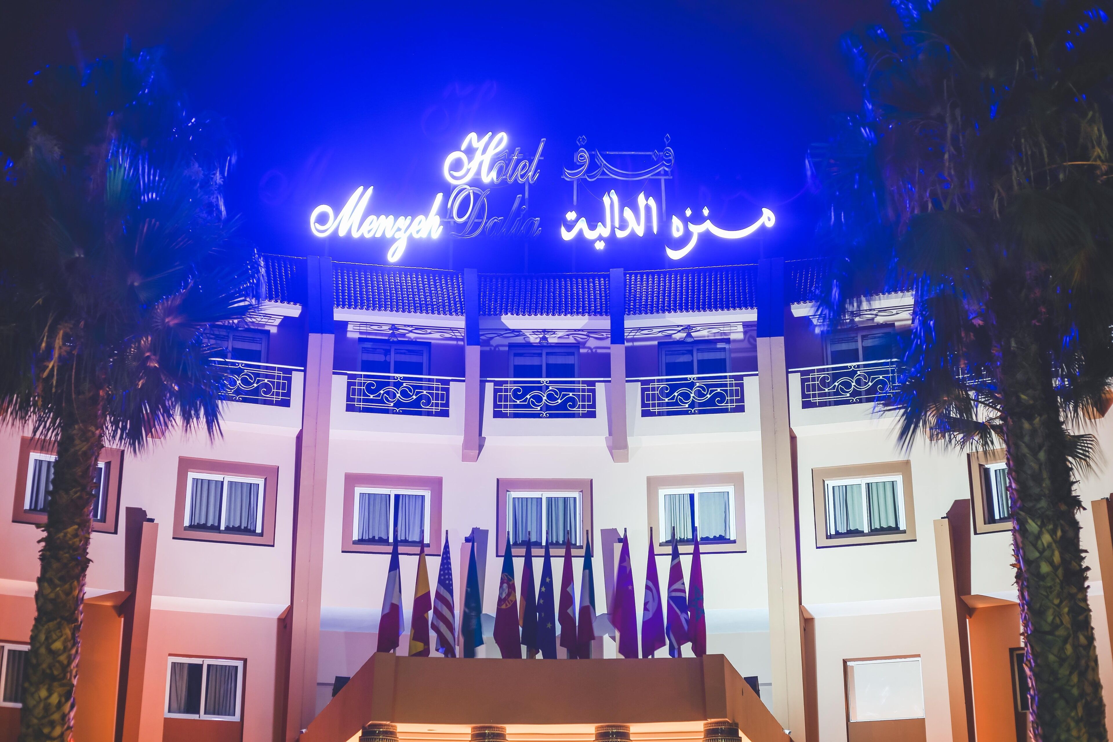 A hotel entrance with a sign in Arabic and English, multiple flags, and a traditional architectural style.