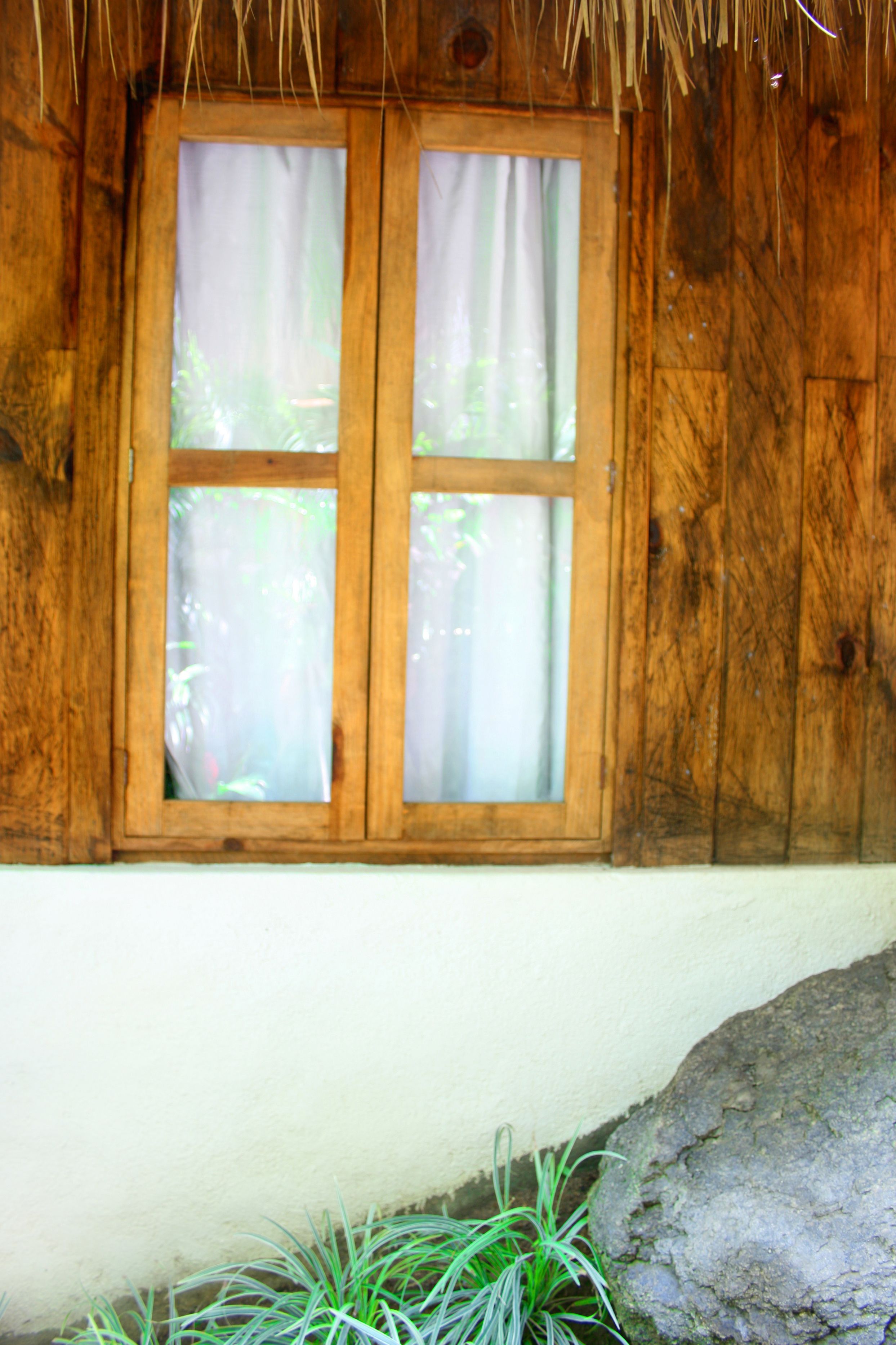 A wooden window with a white curtain, set in a rustic building with a thatched roof.