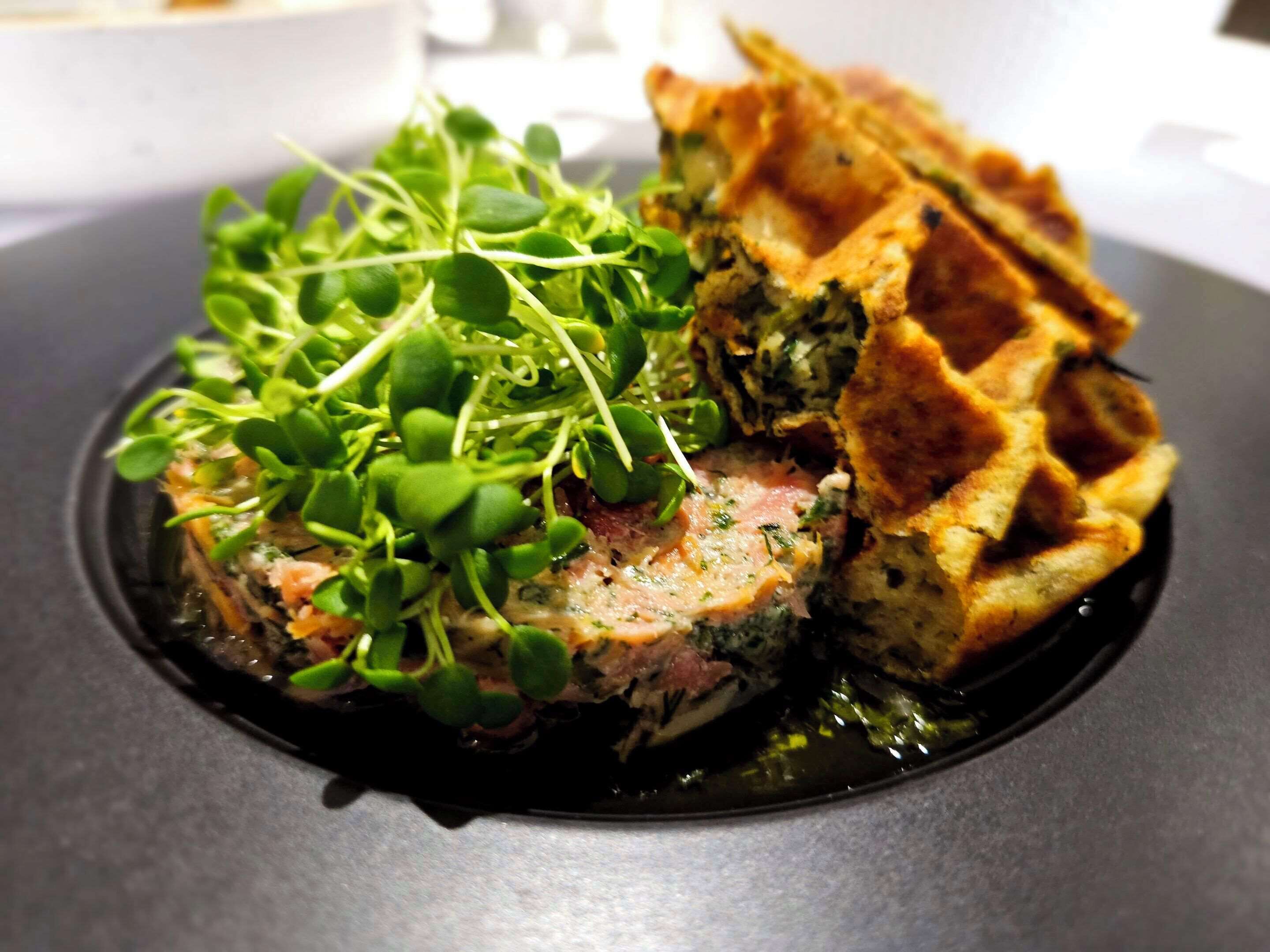 Un plato con una porción de carne coronada con vegetales y un waffle.