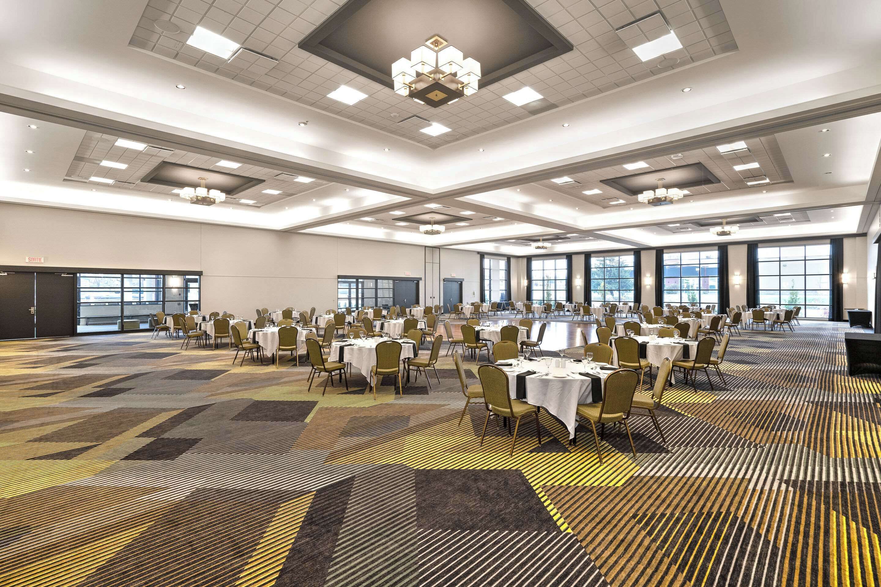 A large banquet hall with multiple round tables and chairs, arranged in rows, with a patterned carpet and a high ceiling with recessed lighting.