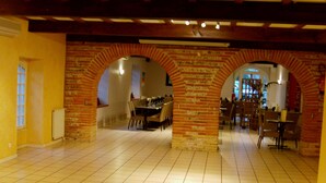 A spacious hall with tiled flooring, brick arches, and wooden beams on the ceiling. Tables and chairs are arranged for dining.