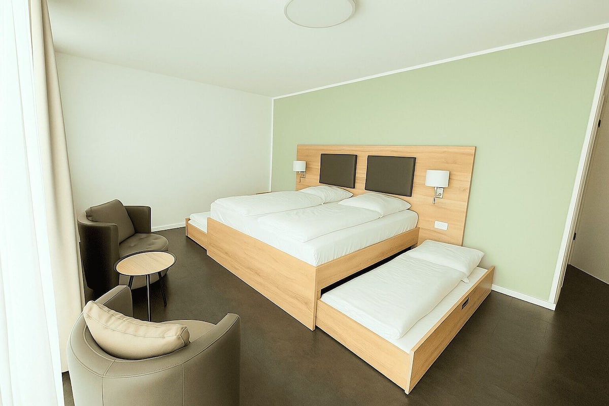 A modern hotel room with a double bed, a small round table, a sofa, and a bedside lamp.