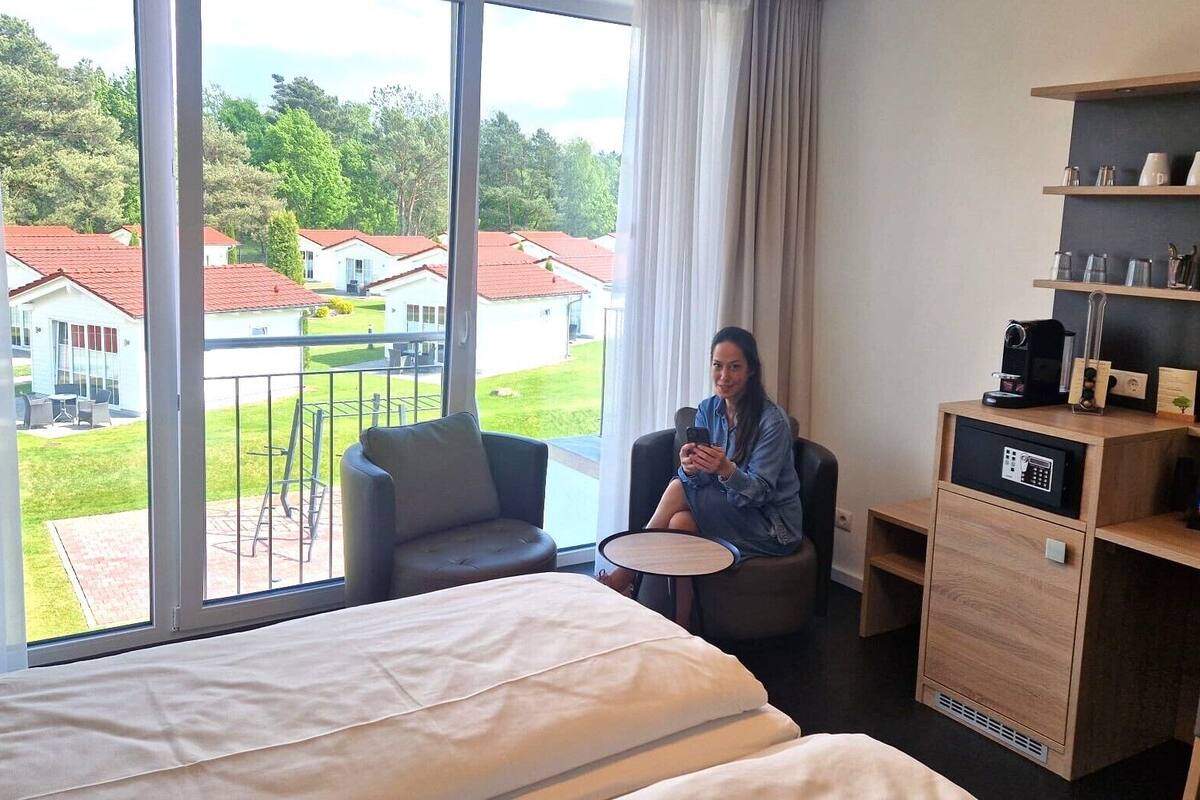 A hotel room with a large bed, a desk, a chair, and a TV. There is a balcony with a view of houses and trees.