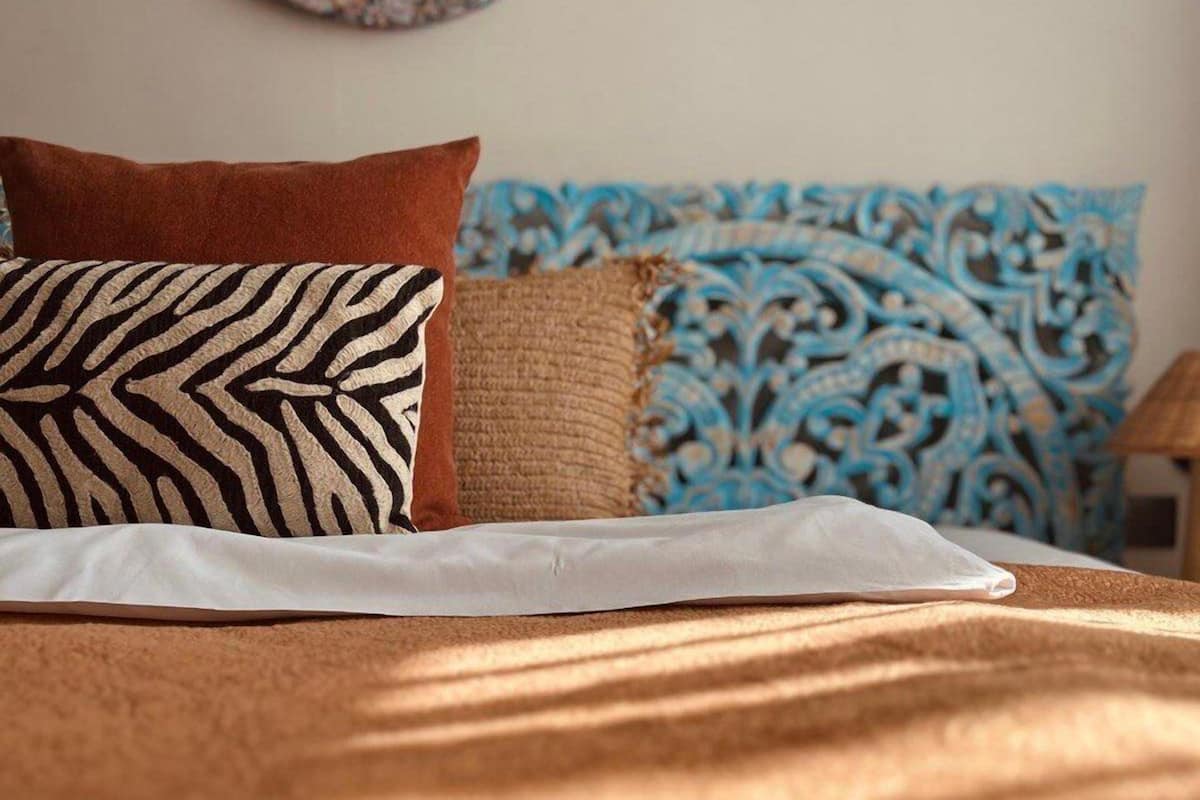 Una cama con sábana blanca, un cojín estampado de cebra, un cojín marrón y un cojín estampado azul.