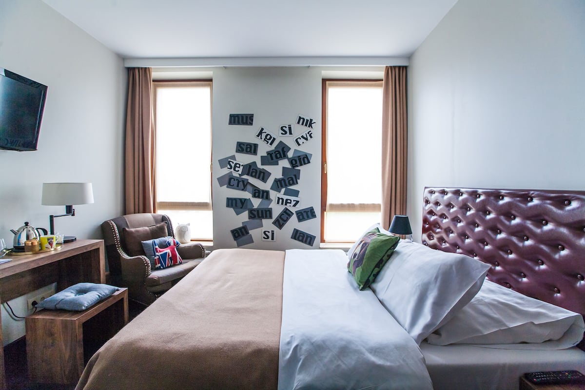 Habitación de hotel con una cama grande, un escritorio con lámpara, una silla y una pared con letras dispersas.