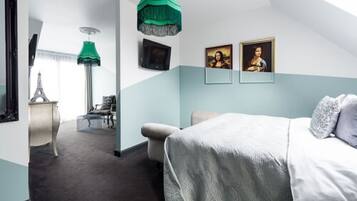 Une chambre d’hôtel avec un lit, un bureau, une chaise et une lampe verte suspendue au plafond. Il y a deux tableaux accrochés au mur.