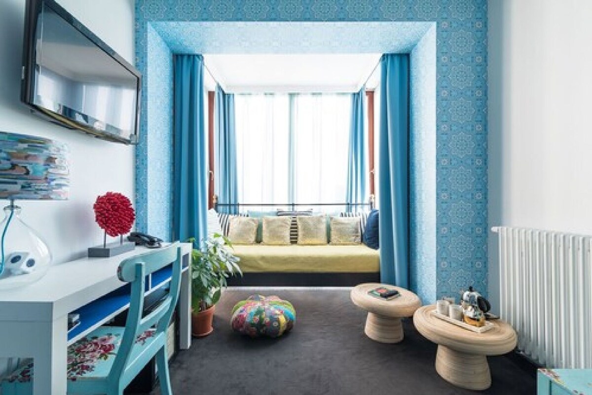 Ein Zimmer mit Bett, Schreibtisch, Stuhl, einem Blumentopf und einem Fenster mit Vorhängen.