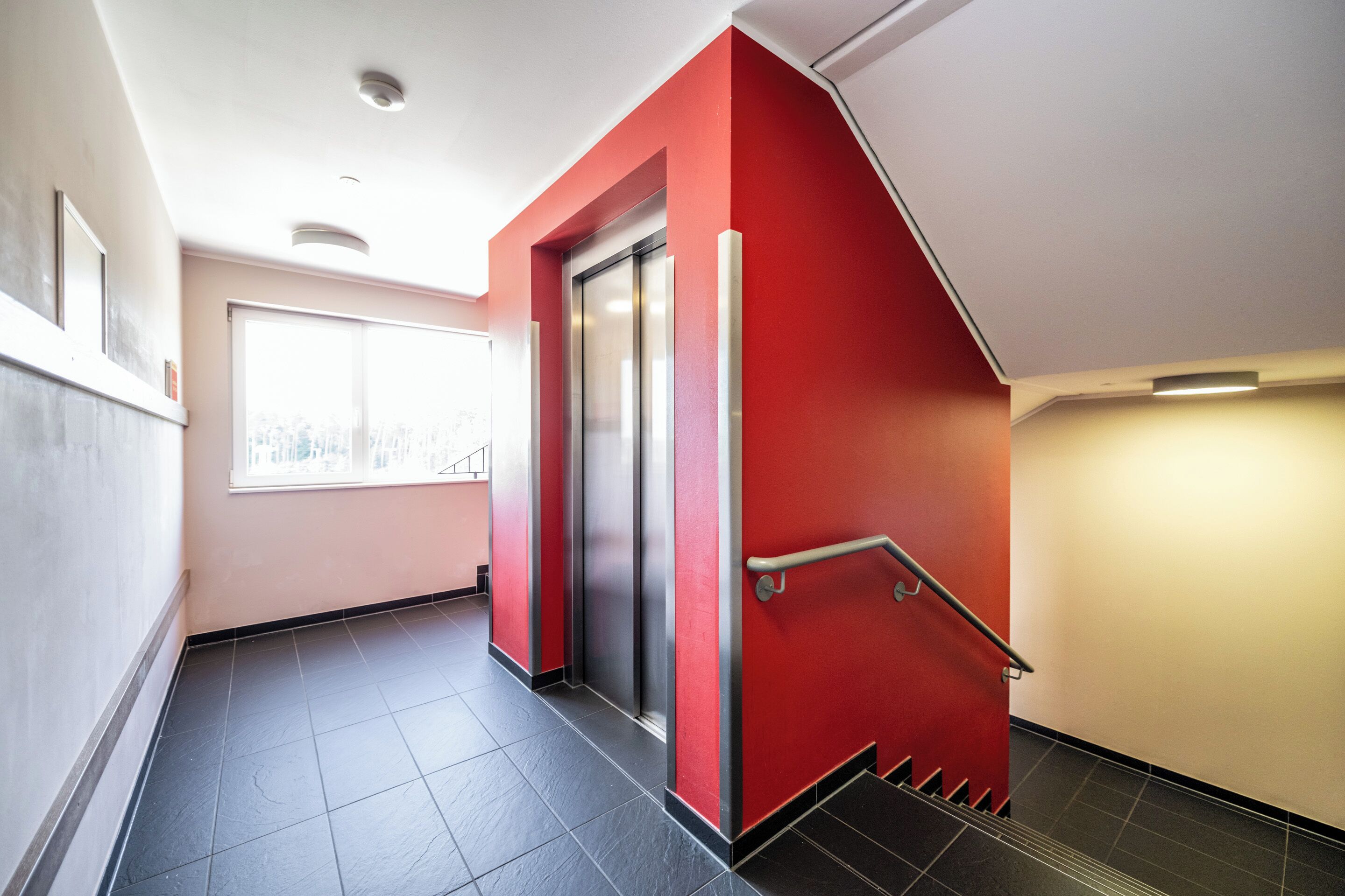 Un pasillo con pared roja, una ventana y una escalera con barandilla.