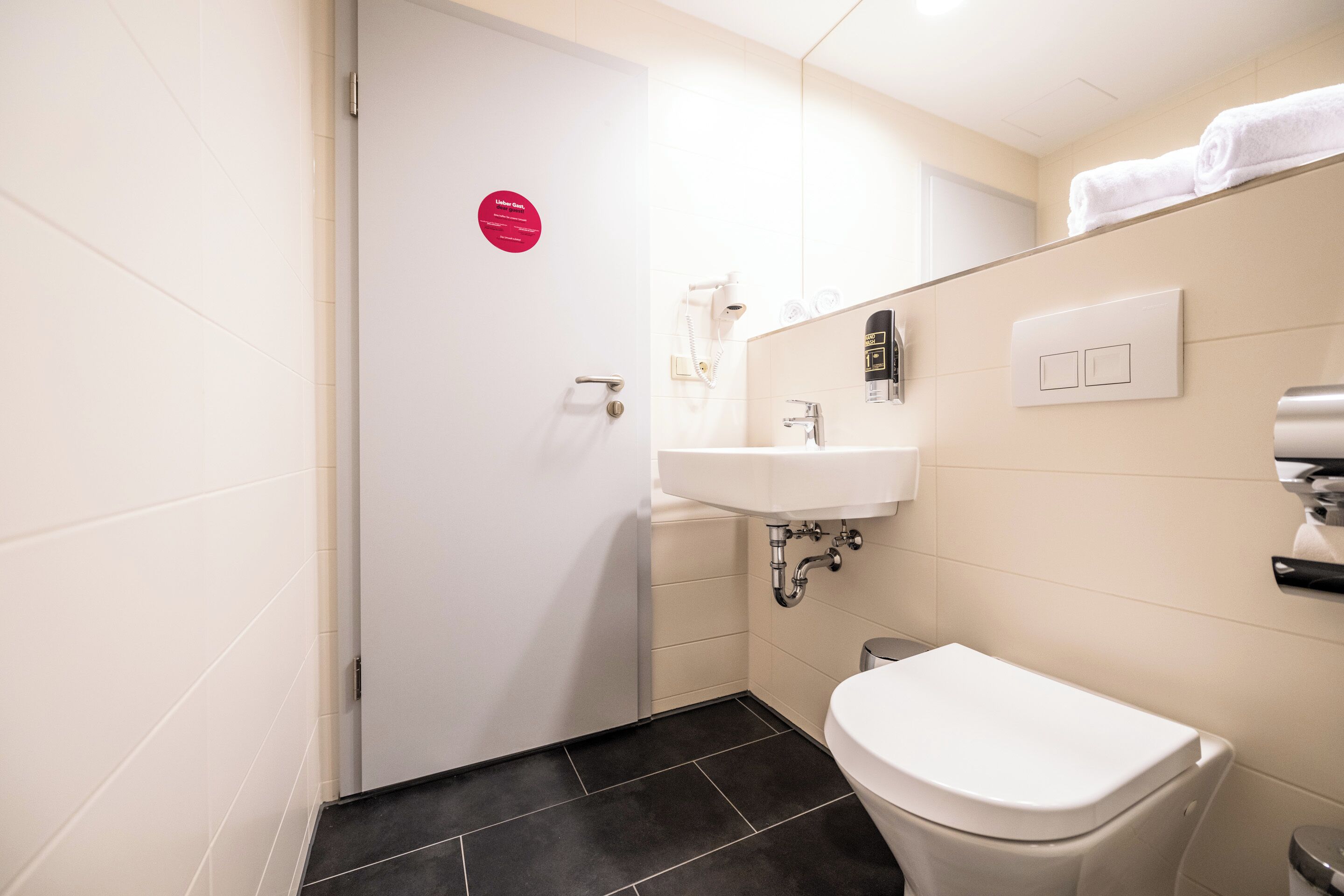 Een badkamer met een wit toilet, een wastafel en een spiegel. Daarboven hangt een handdoekrek bij de wastafel en een deur met een bordje.