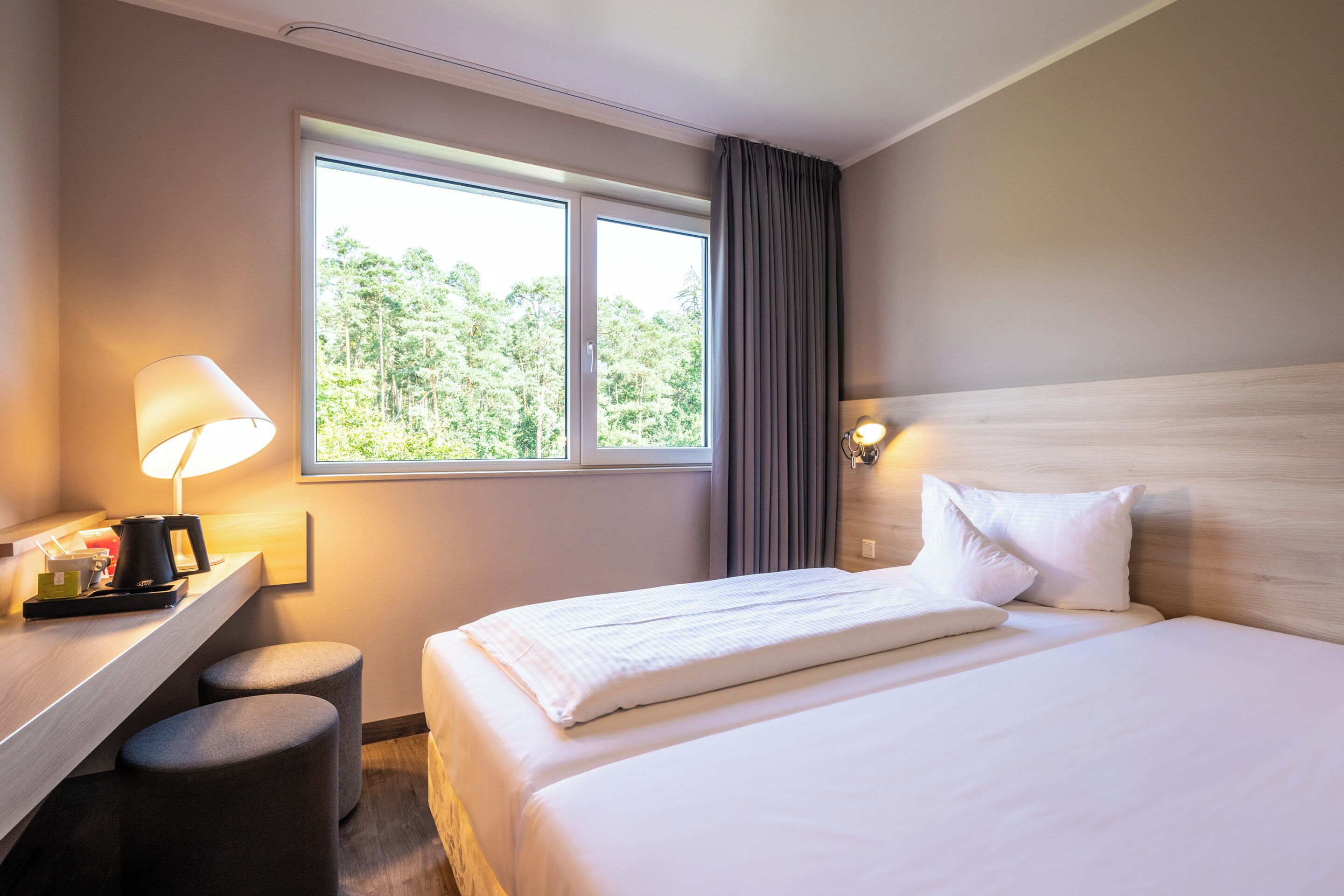 Une chambre d’hôtel avec un lit, un bureau doté d’une lampe, une machine à café et une vue sur la verdure par la fenêtre.