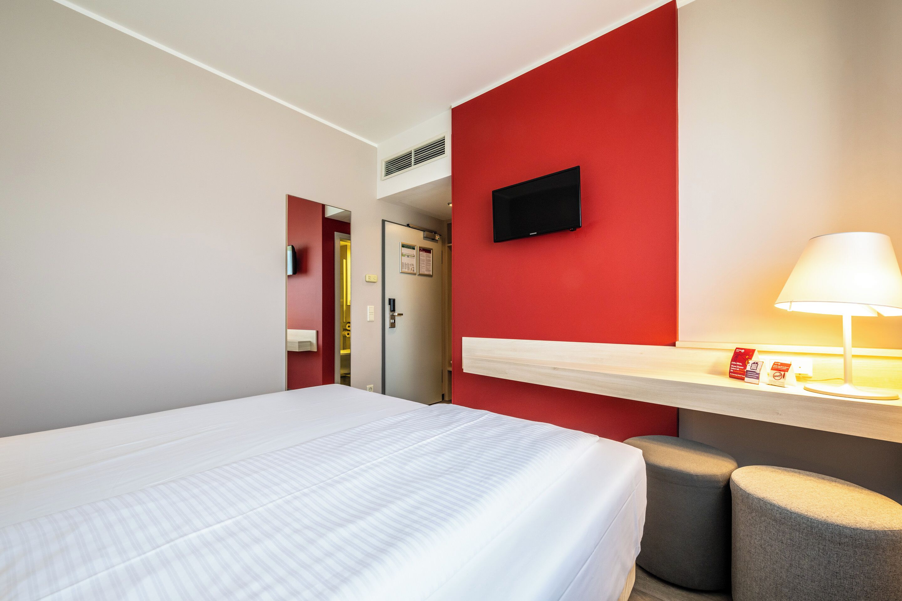 Une chambre d’hôtel avec un lit, un bureau doté d’une lampe et un téléviseur fixé au mur.